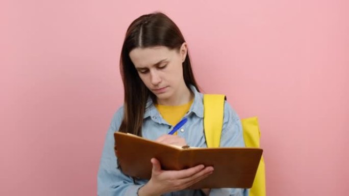 被激怒的少女学生的肖像穿着衬衫和黄色背包在练习册笔记中写下，在工作室的粉红色背景墙上孤立地摆姿势。大