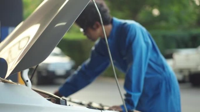 专注于汽车: 亚洲中年汽车修理工穿着蓝色的机械连身裤打开汽车引擎盖，用于检查汽车电池的问题，家庭服务
