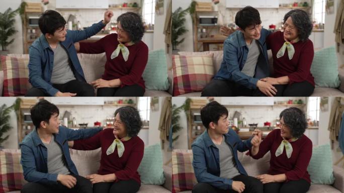 快乐的亚洲成年孙子的慢动作拥抱他的祖母，并在家里的沙发上展示握紧的拳头。男人告诉高级女性，他将在新工