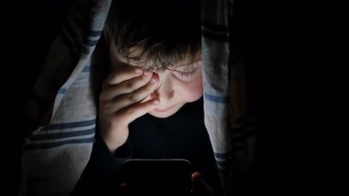 由于屏幕的影响和睡眠的欲望，一个十几岁的男孩在晚上使用智能手机偷偷浏览在线内容，躲在被窝下并用手揉眼