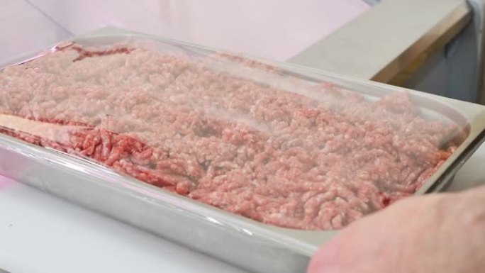 杂货店的肉类销售商将带有食品拉伸膜的新鲜碎肉的托盘关闭，然后将其放在肉类部门的陈列柜中。肉类零售贸易