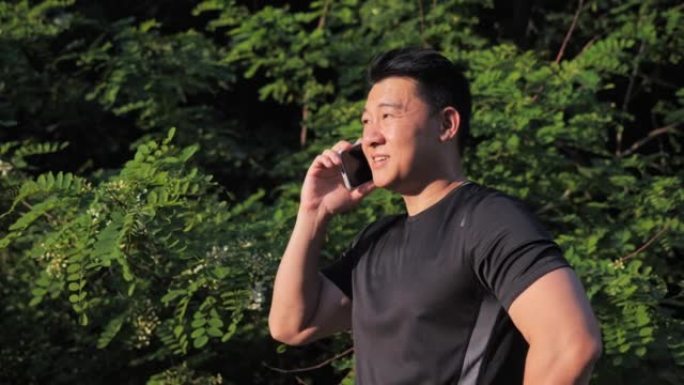 快乐的亚洲男子有手机通话站在阳光下聊天，锻炼后微笑。男跑步者在外面训练。慢跑者分享体育成绩与运动队在