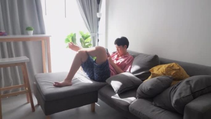 亚洲男孩使用在家中的阳光在窗户附近的沙发上看电影或病毒视频，生活方式的概念。