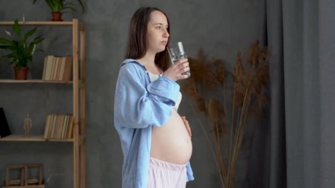 有趣的孕妇在家喝水。年轻的准妈妈想着她的孩子，享受她未来的生活。母亲和怀孕