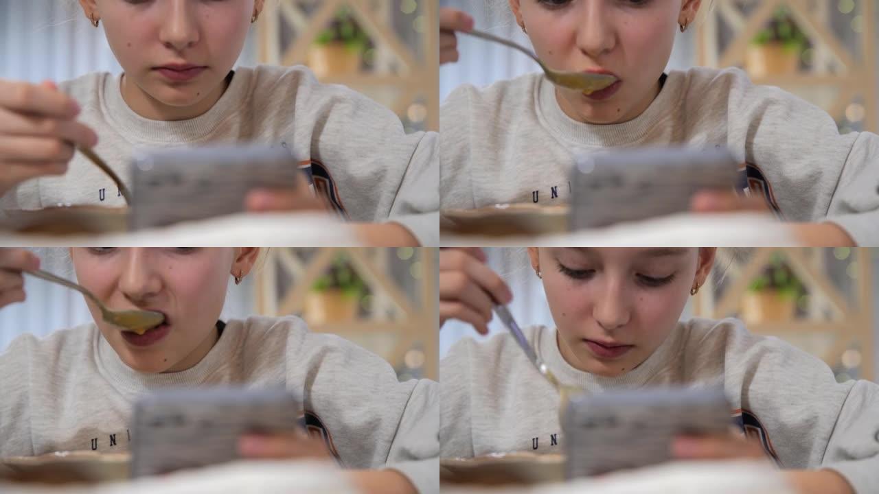 特写饥饿的女孩少年在照明咖啡馆的餐桌上吃奶油汤和油条，同时检查智能手机。坏习惯。