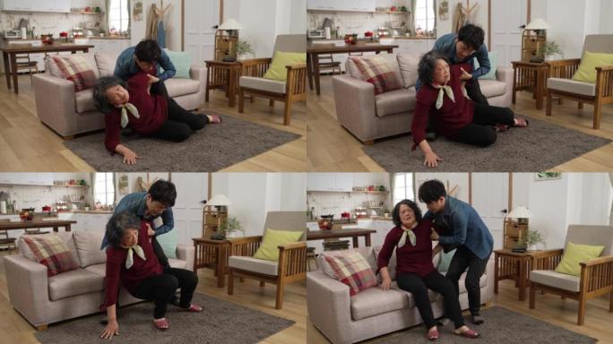 亚洲成年儿子的慢动作在家里摔倒事故后急忙过来帮助他的老母亲坐在沙发上休息。他在她背后放了一个垫子，以