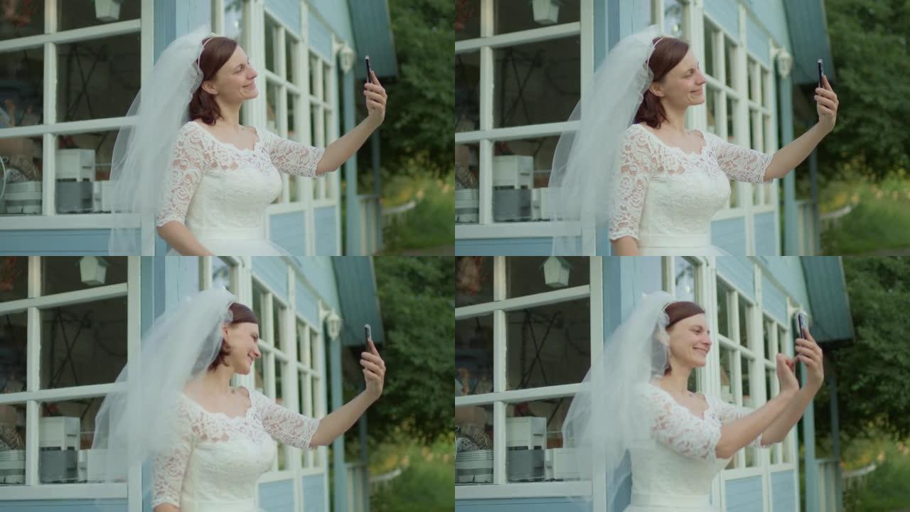 30岁的新娘穿着白色婚纱，用手机站在农村房子附近。拿着手机的新婚快乐。