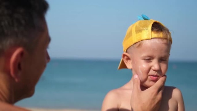 父亲在海滩上给儿子的脸上涂了防护霜。男人的手在孩子的脸上涂防晒霜。海边有防晒霜的可爱小男孩。复制空间