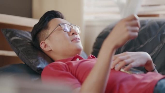 亚洲男子躺在公寓内的沙发上，肚子上放着电话。年轻人在家工作后放松。中国男人在沙发上很舒服