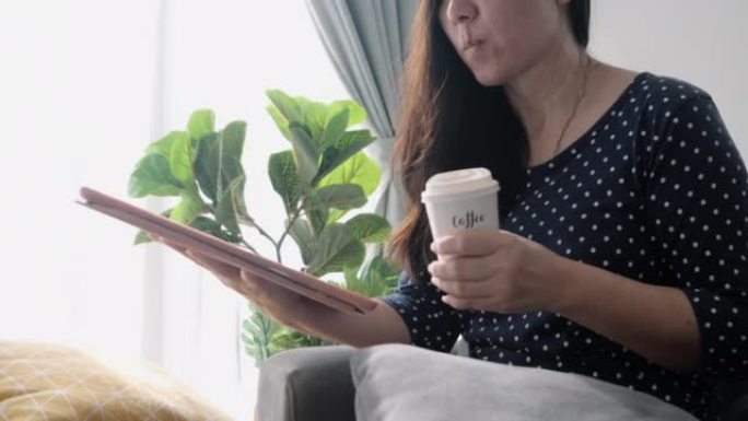亚洲成年女性在沙发上放松，喝一杯咖啡，在家使用靠近窗户的数字平板电脑，生活方式理念。