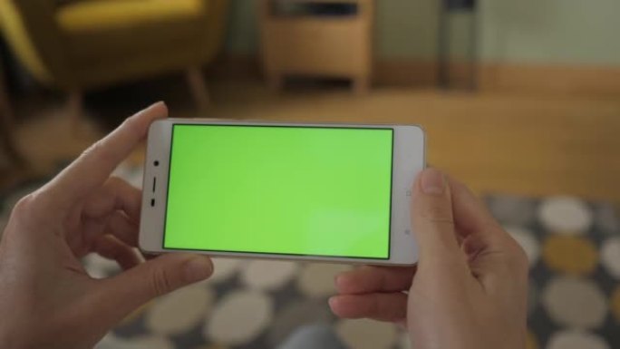 带有绿色模型屏幕色度键的手机上网观看内容视频博客点击中央屏幕。客厅女人的观点坐在椅子上使用带有绿色模