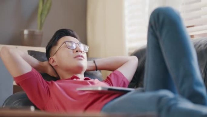 亚洲男子躺在公寓内的沙发上，肚子上放着一台笔记本电脑。年轻人在家工作后放松。中国男人在沙发上很舒服