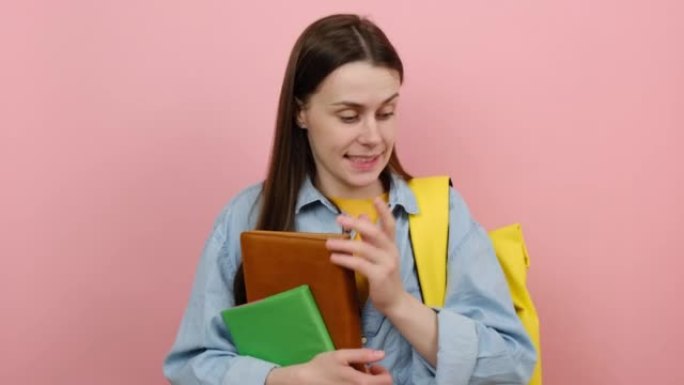 少女学生的肖像穿着衬衫和黄色背包拿着书说哎呀哎呀哦，天哪，在粉红色背景工作室上孤立摆姿势。高中教育大
