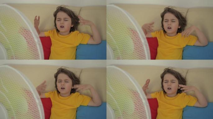 男孩坐在风扇热天的沙发上。夏天炎热时，孩子在家享受电风扇带来的凉风。使用电风扇冷却器冷却自己的通风机