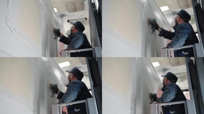 在使用机械砂光机完成喷漆之前，呼吸器中的油漆线工人在汽车墙壁上打磨特写镜头。货车厂油漆车间的油漆工的