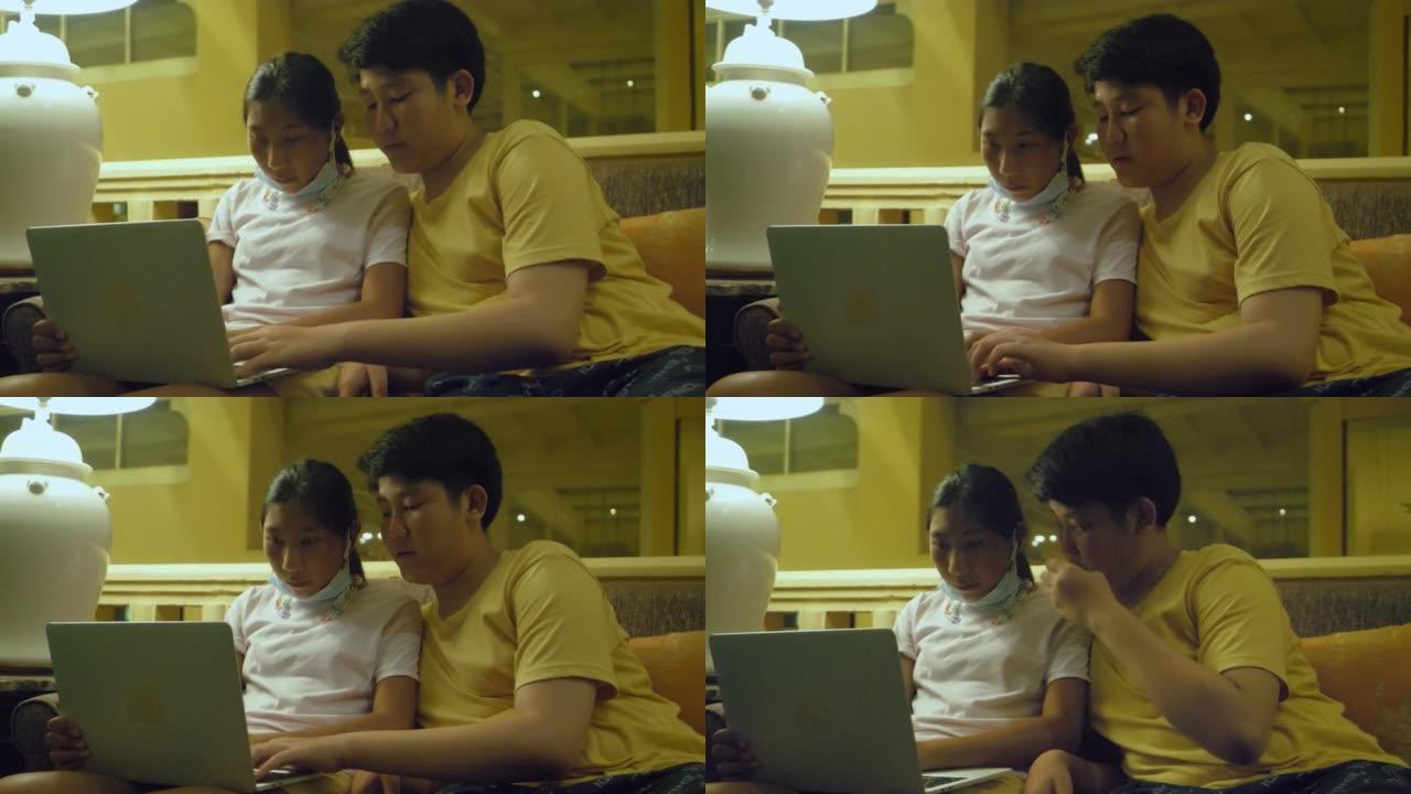 亚洲男孩在度假期间在酒店大堂的沙发上教他的妹妹使用笔记本电脑。