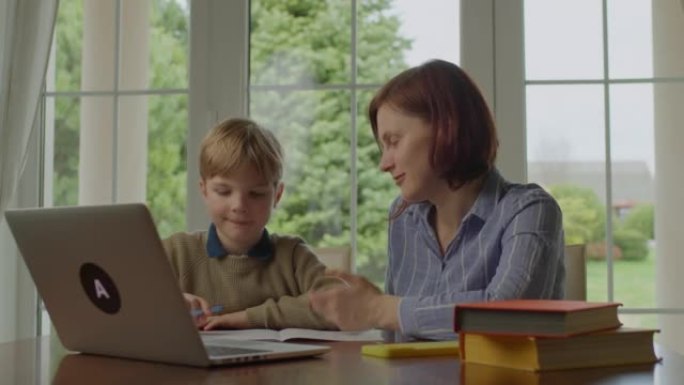 30多岁的年轻母亲在家和小学生做在线作业。家庭使用笔记本电脑一起学习。在线教育过程。