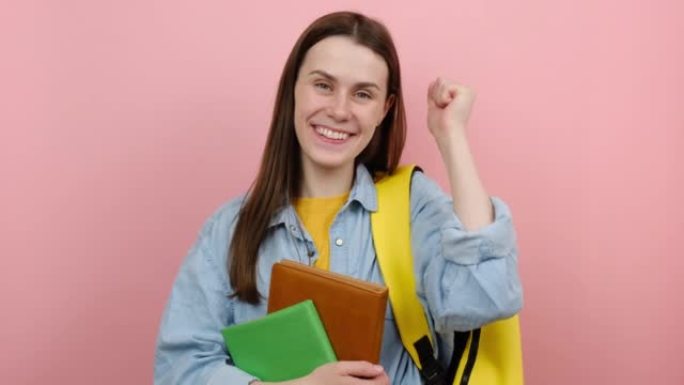 开朗可爱的女孩青少年学生20多岁穿着衬衫和黄色背包握紧拳头，在柔和的粉红色背景工作室中孤立地摆姿势。