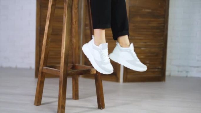 穿着黑色牛仔裤的模特来到木凳上，坐在上面，展示白色新运动鞋。女孩挥动双腿展示时髦的鞋类。