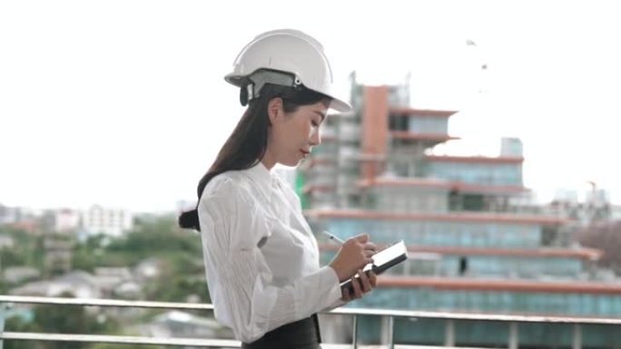 年轻的亚洲女工程师戴着安全帽，在平板电脑上做笔记，同时看着一栋未完成的建筑。