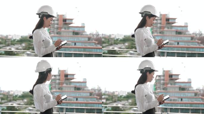 年轻的亚洲女工程师戴着安全帽，在平板电脑上做笔记，同时看着一栋未完成的建筑。