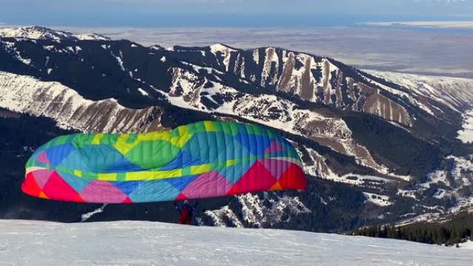 滑翔伞在覆盖着针叶林的雪白斜坡的背景下，随风向上奔跑并抬起滑翔伞的翅膀。