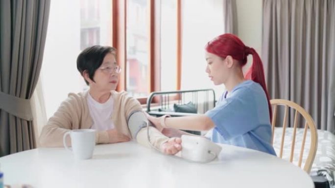 护士通过使用自动血压监测器对成熟的亚洲高级女性进行血压测量。照顾者在家探望。家庭保健和疗养院概念。实