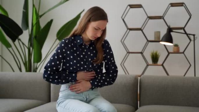一名年轻女子正坐在家里的沙发或沙发上，腹痛严重。床上有经前期、经期或中毒的女人，黑发女人患有腹部绞痛