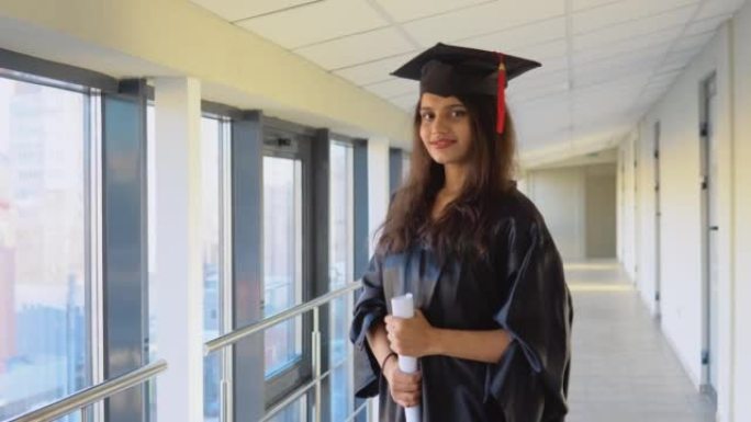 披风的巴基斯坦女毕业生手里拿着文凭站着，微笑着
