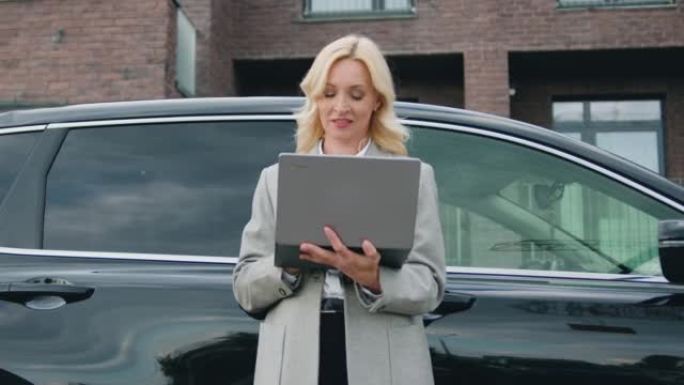 迷人的金发女司机站在她的车旁，在城市街道上使用笔记本电脑。阅读新消息或浏览视频或照片或社交网络时微笑