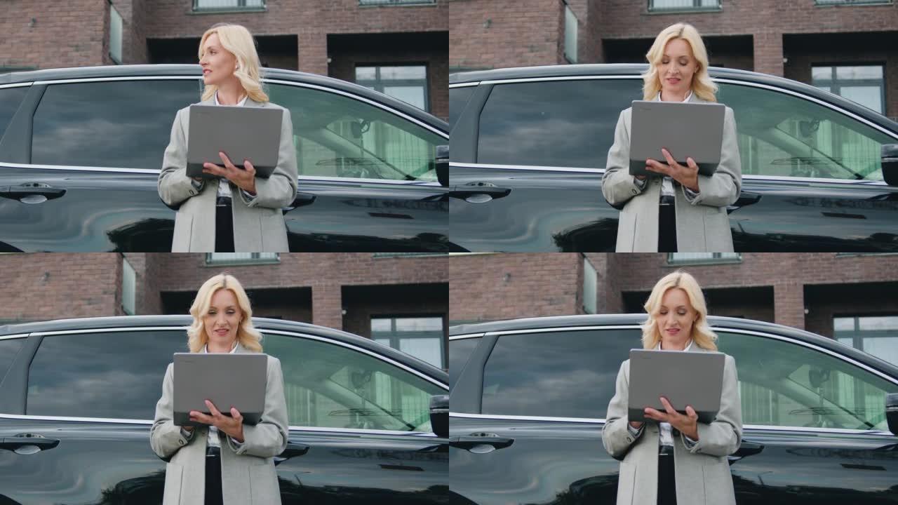 迷人的金发女司机站在她的车旁，在城市街道上使用笔记本电脑。阅读新消息或浏览视频或照片或社交网络时微笑