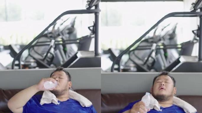 精疲力竭的胖子坐在沙发上喝塑料瓶里的冷水，经过艰苦的锻炼后感到放松。