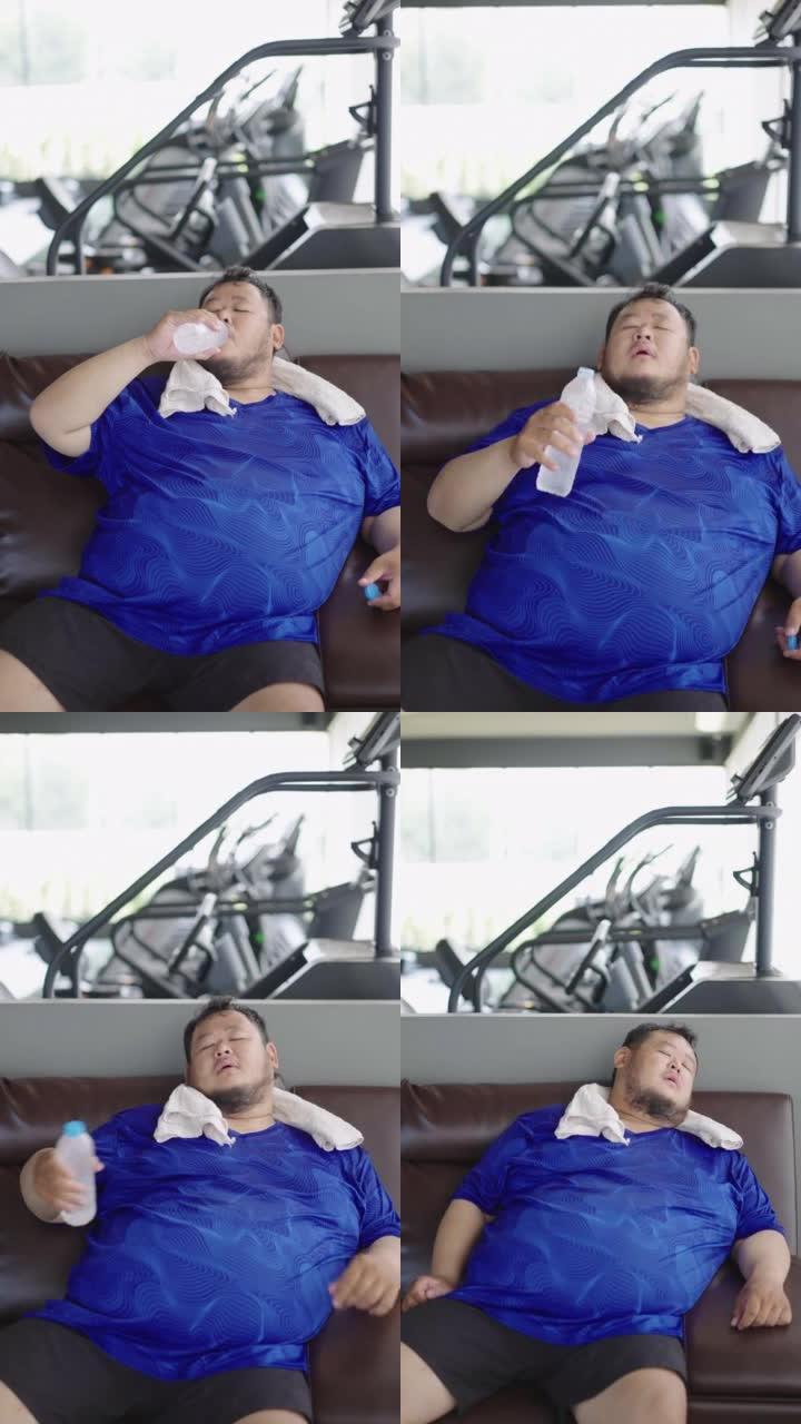 精疲力竭的胖子坐在沙发上喝塑料瓶里的冷水，经过艰苦的锻炼后感到放松。