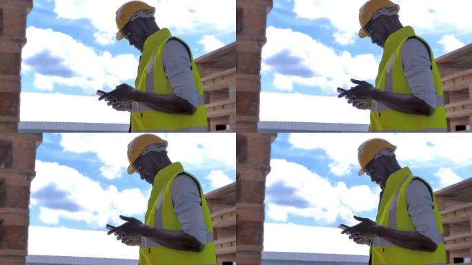 一名戴着防护头盔并穿着制服的专业男性建筑工人正在使用智能手机在建筑工地背景下工作。工业和施工现场概念