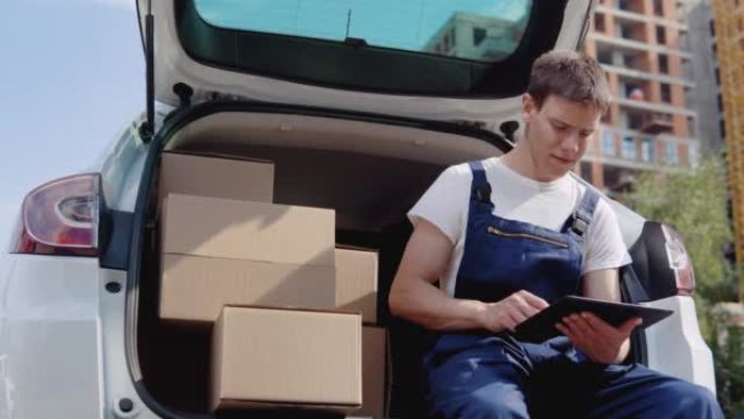 快递员坐在打开的后备箱边缘，装满了一箱货物，并填写了文件