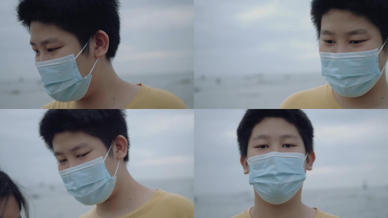 亚洲男孩戴着口罩站在海滩上，日出之前看着相机，这是新的常态和社交距离的生活方式概念。