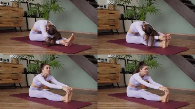年轻女子在家伸展。一个穿着紫色西装的女人坐在瑜伽垫上，弯腰弯腰，将脚拉向自己，伸展腿部肌肉