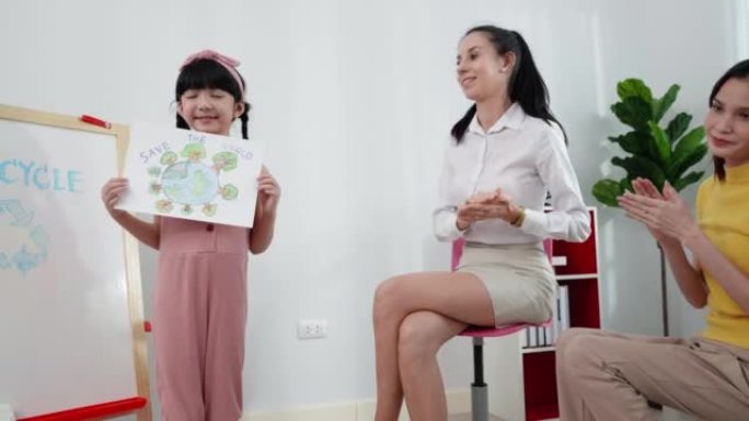 可爱的亚洲六岁女孩站在教室前，以拯救世界为主题展示她的彩色铅笔画，她用很多树木画了全球封面，而老师和
