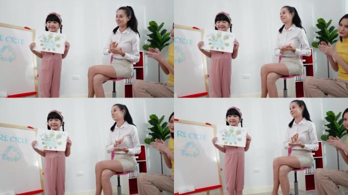 可爱的亚洲六岁女孩站在教室前，以拯救世界为主题展示她的彩色铅笔画，她用很多树木画了全球封面，而老师和