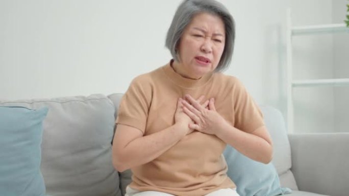 亚洲老年妇女坐在沙发上，双手放在她的胸部中央，因为她感到胸闷，在她的客厅里，她的胸部有灼痛。