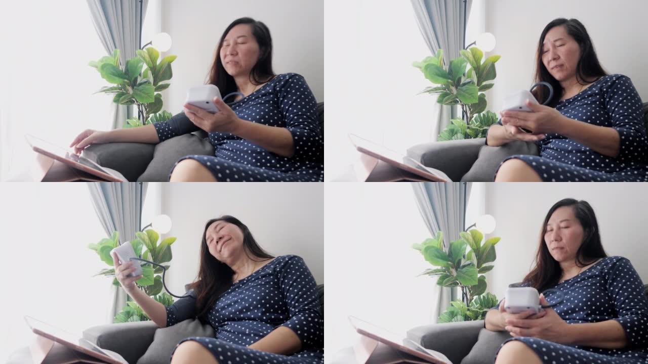 亚洲妇女与血压测试和寻找数字表在网站上的建议，而坐在沙发上靠窗在家，健康的生活方式的概念。
