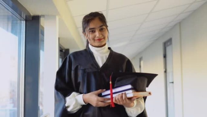 披风的巴基斯坦女毕业生手里拿着书站着，微笑着
