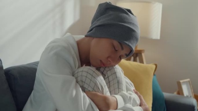 绝望的年轻亚洲癌症患者坐在家里客厅的沙发上。