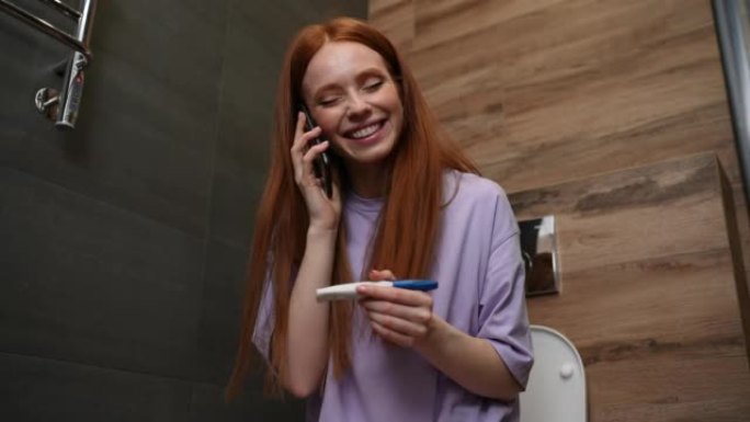 开朗的年轻女子在怀孕测试中保持阳性结果，并通过智能手机与丈夫坐在家里浴室的马桶上交谈。
