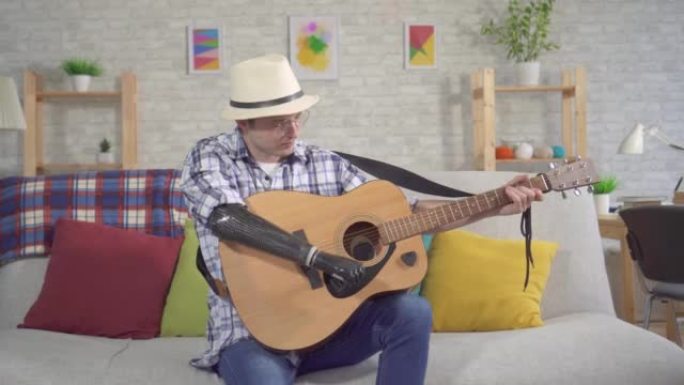 戴着仿生假肢帽子的年轻人坐在客厅的沙发上弹奏原声吉他