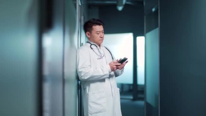 亚洲精疲力竭的医生在现代医院诊所的走廊休息时站着时使用手机智能手机。