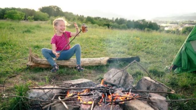 小女孩意外地把香肠棒掉进了篝火，有趣地笑了起来，坐在帐篷营地的木头上。家庭户外野餐活动概念4k镜头。