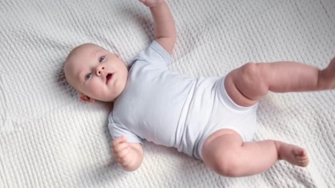 美丽的新生儿躺在白色的毯子上。小孩穿着白色裤子和蓝色紧身衣。可爱的小宝贝正看着镜头做手势。童年，健康