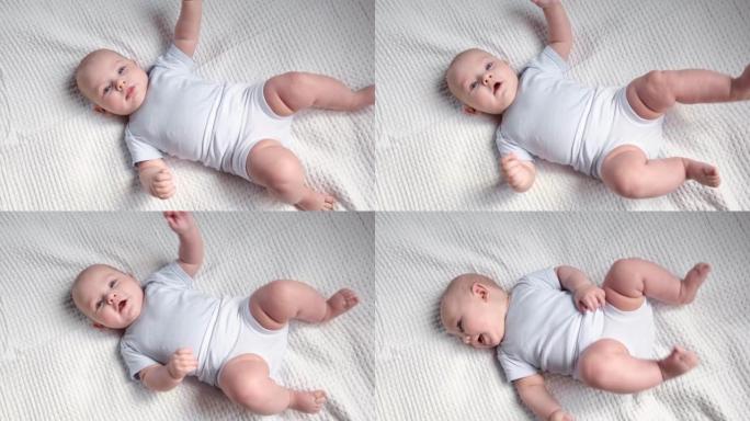 美丽的新生儿躺在白色的毯子上。小孩穿着白色裤子和蓝色紧身衣。可爱的小宝贝正看着镜头做手势。童年，健康
