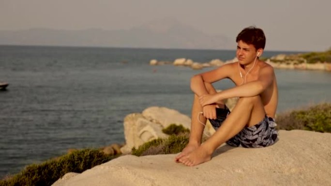 白人十几岁的男孩坐在海边的岩石上听音乐